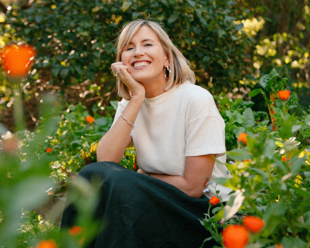 Ruby Jones sits amongst orange flowers resting her chin in her hand and smiling at the camera
